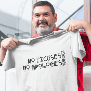 Man in his 50s with a white beard, joyfully holding up a vanilla-colored t-shirt featuring the empowering quote "No Excuses No Apologies No Regrets", showcasing a positive attitude and an inspirational message, perfect for expressing determination and resilience in life.